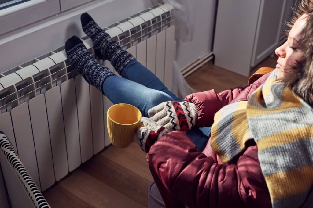 A woman in a coat and gloves warming her feet on a radiator